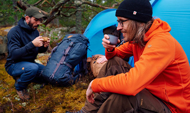 Zwei Personen sitzen neben einem blauen Zelt im Wald. Einer trägt eine orangefarbene Jacke und trinkt einen Schluck. Der andere, eine dunkle Jacke und eine Mütze, bereitet Essen zu. Rucksäcke und Campingausrüstung liegen in der Nähe auf dem moosigen Boden.
