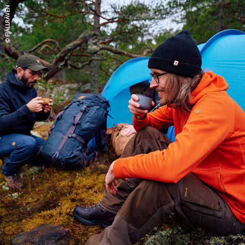Zwei Personen campen im Wald. Einer trägt eine orangefarbene Jacke und eine schwarze Mütze, sitzt da und hält eine Tasse in der Hand. Der andere trägt eine dunkle Jacke und eine Mütze und sitzt daneben. Im Hintergrund sind ein blaues Zelt und Rucksäcke zu sehen, umgeben von Bäumen.