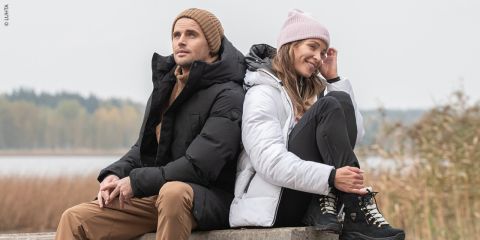 Ein Mann und eine Frau sitzen im Freien auf einem Betonvorsprung. Sie tragen Winterkleidung. Der Mann in schwarzer Jacke und Mütze blickt zur Seite, die Frau in weißer Jacke und rosa Mütze lächelt und blickt geradeaus. Im Hintergrund sind ein Gewässer und Schilf zu sehen.