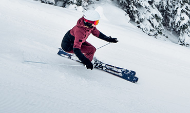 Ein Skifahrer in rot-schwarzer Kleidung und weißem Helm fährt einen schneebedeckten Hang hinunter. Er nutzt Stöcke, um das Gleichgewicht zu halten. Im Hintergrund sind schneebedeckte Bäume zu sehen.