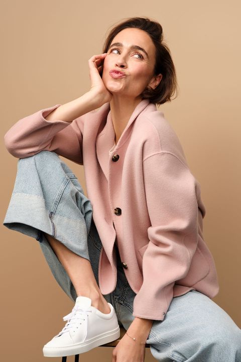 Eine Frau in rosa Blazer und Jeans sitzt verspielt auf einem Hocker und stützt ihr Kinn auf ihre Hand. Sie trägt weiße Turnschuhe, blickt leicht nach oben und schmollt vor einem beigen Hintergrund.