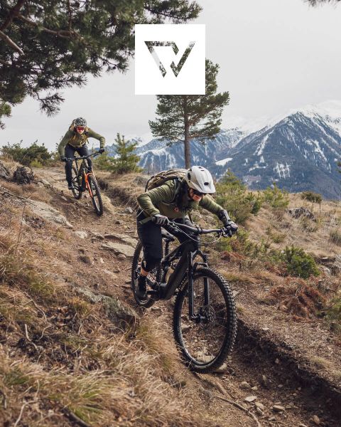 Zwei Radfahrer fahren mit Mountainbikes auf einem schroffen Pfad in einer Bergregion mit schneebedeckten Gipfeln im Hintergrund. Sie tragen Helme und Outdoor-Ausrüstung. Oben im Bild ist ein Logo zu sehen.