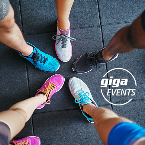 Fünf Personen stehen im Kreis, die Füße zeigen nach innen. Sie tragen bunte Sportschuhe. Der Hintergrund ist ein dunkel strukturierter Boden. Auf der rechten Seite befindet sich das Logo „giga EVENTS“.
