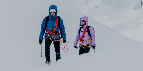 Zwei Personen in Winterkleidung wandern durch den Schnee. Sie tragen Helme und Klettergurte. Sie sind durch ein rotes Seil verbunden und tragen Eispickel. Im Hintergrund ist eine verschneite Berglandschaft zu sehen.