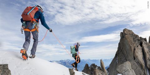 Zwei Bergsteiger überqueren einen schneebedeckten Bergkamm, aneinander geseilt. Sie tragen Rucksäcke und Steigeisen, der Himmel ist teilweise bewölkt. Im Hintergrund sind felsige Gipfel zu sehen, bis zum Horizont erstreckt sich eine Bergkette.