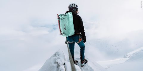 Ein Bergsteiger mit Helm und großem grünen Rucksack steht auf einem schmalen, schneebedeckten Grat. Der Himmel ist bedeckt, Nebel und Wolken verhüllen die Landschaft, was eine dramatische und abenteuerliche Atmosphäre schafft.