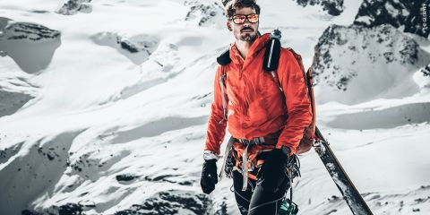 Ein Mann in roter Jacke und Skiausrüstung steht mit Skiern in der Hand auf einem schneebedeckten Berg. Er trägt eine Sonnenbrille und Handschuhe. Im Hintergrund sind Klippen und schneebedeckte Gipfel zu sehen.