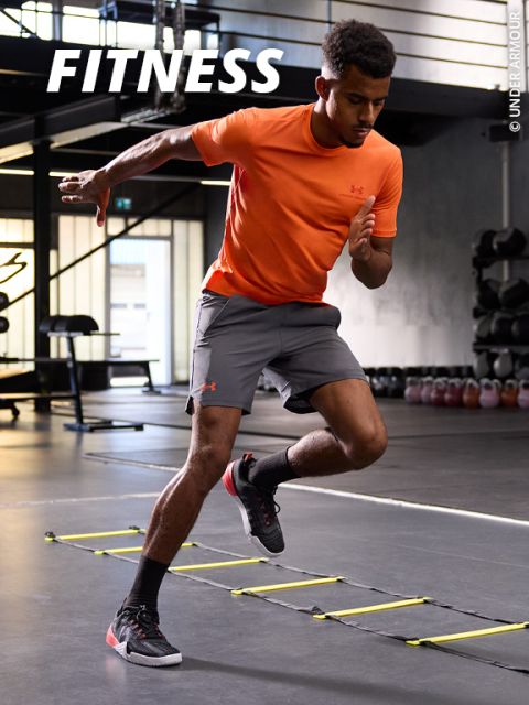 Mann in orangefarbenem Hemd und grauen Shorts macht Koordinationsübungen mit der Leiter in einem Fitnessstudio. Oben steht das Wort „FITNESS“.