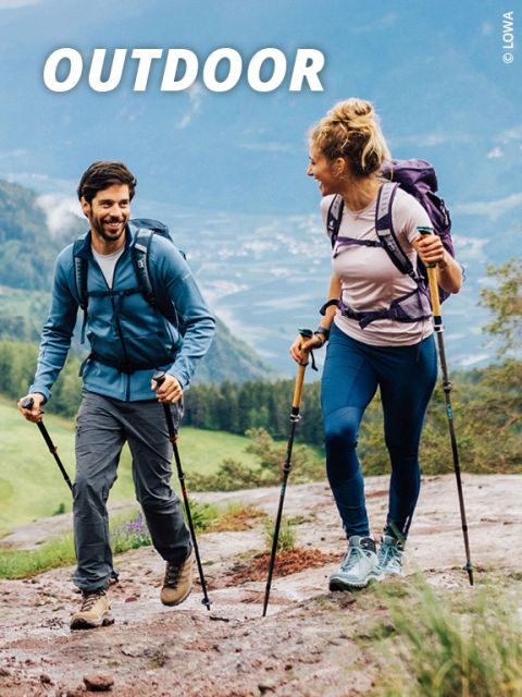 Ein Mann und eine Frau wandern mit Trekkingstöcken und Rucksäcken auf einem steinigen Pfad. Sie lächeln sich an, im Hintergrund ein malerischer Blick auf Hügel und ein Tal. Oben im Bild steht das Wort „OUTDOOR“.