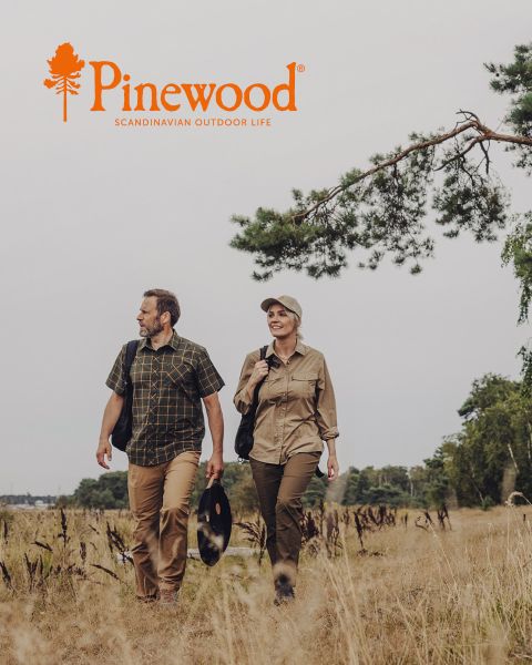 Ein Mann und eine Frau in Outdoor-Kleidung gehen unter einem bewölkten Himmel durch eine Wiese. Die Frau trägt eine schwarze Tasche, während der Mann einen Hut hält. Über ihnen sind das Pinewood-Logo und der Text „Scandinavian Outdoor Life“ zu sehen.
