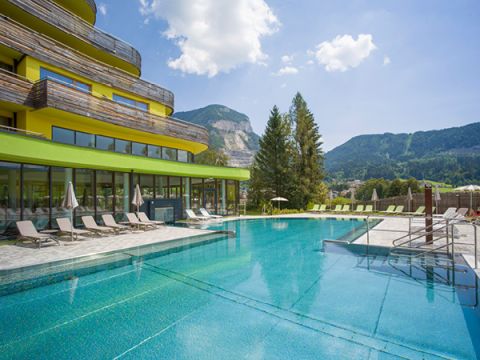Modernes Hotel mit leuchtend grünen Akzenten und geschwungener Architektur neben einem Außenpool. Rund um den Pool stehen Liegestühle und Sonnenschirme bereit. Im Hintergrund üppig grüne Berge und ein teilweise bewölkter Himmel.