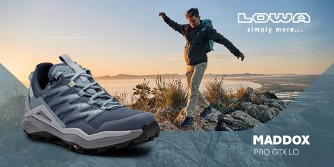 Ein Mann wandert bei Sonnenuntergang mit ausgestreckten Armen durch felsiges Gelände. Im Vordergrund ist eine Nahaufnahme eines marineblauen Lowa Maddox Pro GTX Lo SL Wanderschuhs zu sehen. Der Text lautet: „Lowa einfach mehr... MADDOX PRO GTX LO SL.“.