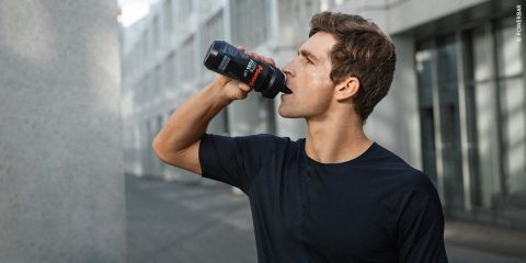 Eine Person in einem schwarzen Hemd trinkt im Freien aus einer Sportflasche. Sie steht auf einem gepflasterten Weg zwischen modernen Gebäuden und hat den Kopf zum Trinken leicht in den Nacken gelegt.