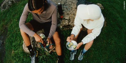 Zwei Personen sitzen draußen auf einem Felsen und genießen eine Mahlzeit. Eine Person trägt ein Langarmshirt und Shorts, die andere einen Kapuzenpullover und Shorts. Beide haben Metallschüsseln mit Gurkenscheiben in der Hand. Eine Thermoskanne und ein Apfel stehen daneben im Gras.