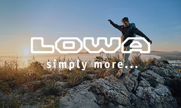 Eine Person in Outdoor-Kleidung balanciert bei Sonnenaufgang über felsigem Gelände und blickt auf eine malerische Landschaft. Der Text „LOWA simply more…“ ist über dem Bild zu sehen.