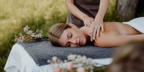 Eine Frau liegt im Freien auf einer Massageliege und genießt eine Schultermassage. Sie ist von viel Grün umgeben, und rund um die Liege stehen Blumen, die eine ruhige und entspannende Atmosphäre schaffen.