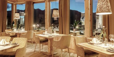 Ein gemütliches Restaurant mit Holztischen und gemusterten Stühlen. Große Fenster bieten einen Blick auf die Berge in der Abenddämmerung. Warmes Licht von Hängelampen schafft eine einladende Atmosphäre, und jeder Tisch ist elegant gedeckt.
