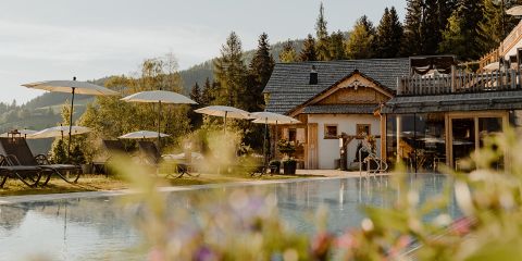 Ein ruhiger Außenpool, umgeben von Liegestühlen und Sonnenschirmen, inmitten üppiger Vegetation und einer rustikalen Holzhütte. Hohe Kiefern und ferne Hügel sind unter einem klaren Himmel sichtbar und schaffen eine ruhige, idyllische Atmosphäre.
