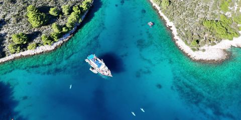 Luftaufnahme eines Bootes mit blauen Akzenten, das in einer türkisfarbenen Bucht vor Anker liegt, umgeben von üppigen grünen Bäumen an felsigen Ufern. Mehrere kleine Kajaks treiben in der Nähe auf dem klaren, blauen Wasser.