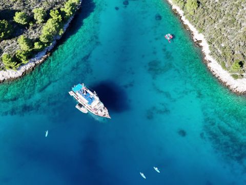 Luftaufnahme eines Bootes mit blauen Akzenten, das in einer türkisfarbenen Bucht vor Anker liegt, umgeben von üppigen grünen Bäumen an felsigen Ufern. Mehrere kleine Kajaks treiben in der Nähe auf dem klaren, blauen Wasser.