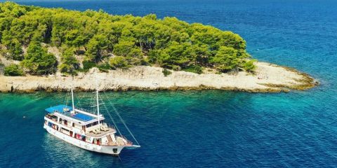 Eine große weiße Yacht mit mehreren Decks liegt vor Anker in der Nähe einer üppig grünen Insel mit felsiger Küste. Das klare blaue Wasser umgibt die Insel, und der Himmel ist hell. Die Szenerie ist ruhig und einladend.