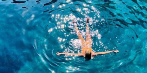 Eine Person schwimmt auf dem Rücken im klaren, blauen Wasser und erzeugt dabei sanfte Wellen. Das Sonnenlicht wird von der Oberfläche reflektiert und erzeugt einen glitzernden Effekt. Die Arme sind ausgestreckt, und die Person wirkt entspannt.