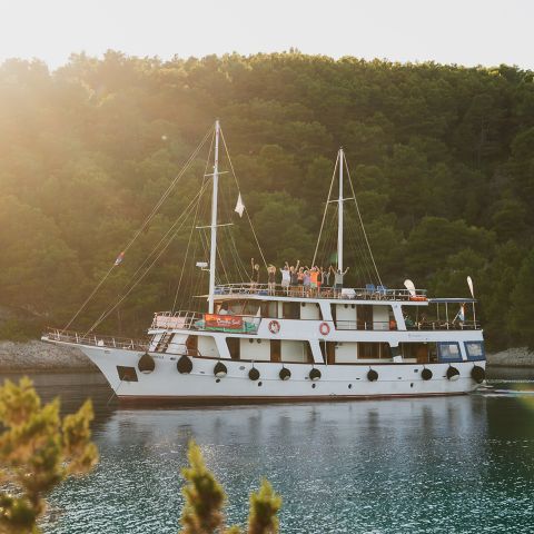 Eine große weiße Yacht mit zwei Masten liegt nahe einer bewaldeten Küste vor Anker. Mehrere Personen winken auf dem Oberdeck. Die Sonne geht unter und taucht die Szene in ein warmes, goldenes Licht.