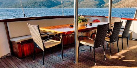 Auf einem Holzdeck eines Bootes befindet sich ein Essbereich mit einem polierten Holztisch, umgeben von gepolsterten Stühlen und einer eingebauten Bank. Ein Blumengesteck schmückt den Tisch. Im Hintergrund sind ein ruhiges Meer und baumbedeckte Hügel unter blauem Himmel zu sehen.