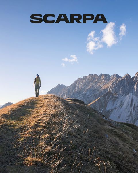 Person, die unter einem klaren blauen Himmel auf einem grasbewachsenen Bergrücken mit schroffen Bergen im Hintergrund wandert, oben das „Scarpa“-Logo.