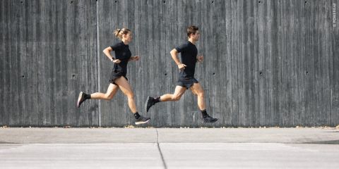 Zwei Personen in Sportkleidung joggen nebeneinander auf einem gepflasterten Gehweg vor einer grauen Betonmauer. Beide tragen schwarze Kleidung und Laufschuhe. Die Szene erinnert an eine urbane Umgebung.