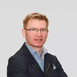 Eine Person mit kurzen, hellbraunen Haaren und Brille, die ein blaues Hemd und einen dunklen Blazer trägt, blickt mit einem leichten Lächeln nach vorne. Der Hintergrund ist schlicht hellgrau.