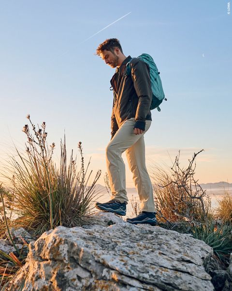 Ein Mann wandert bei Sonnenuntergang in Outdoor-Kleidung und mit Rucksack durch felsiges Gelände. Über dem Bild steht in großem weißen Text: „-15% AUF OUTDOOR-SCHUHE*“.