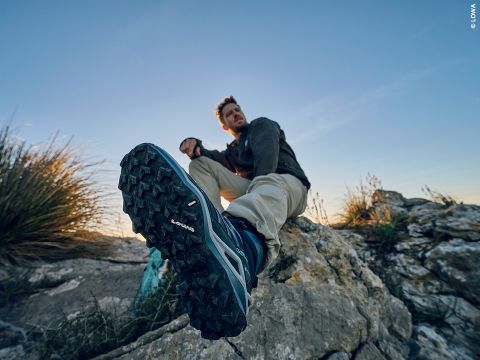 Ein Mann in Outdoor-Kleidung sitzt bei Sonnenuntergang auf einem Felsen. Im Vordergrund ist sein Fuß zu sehen, der die robuste Sohle seines Lowa-Wanderschuhs zeigt. Im Hintergrund sind Gräser und ein klarer Himmel zu sehen.
