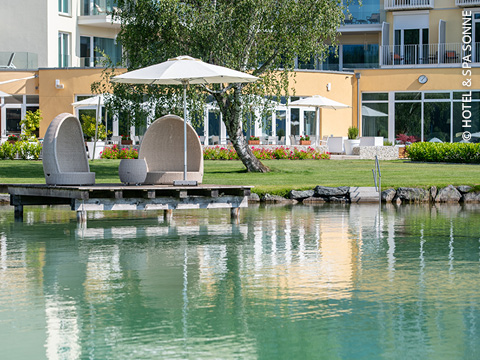 Zwei geflochtene Liegestühle mit weißen Sonnenschirmen stehen auf einem Holzsteg an einem ruhigen See vor einem modernen Hotelgebäude mit großen Fenstern und grünen Rasenflächen.