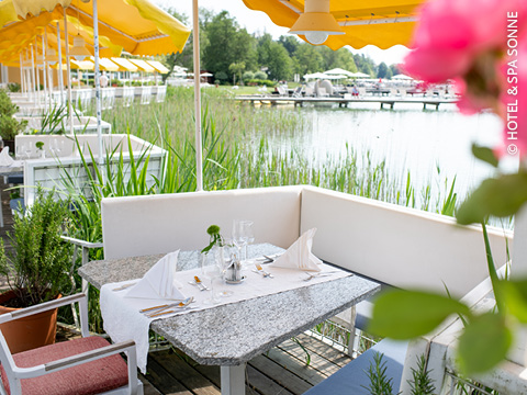 Ein für zwei Personen gedeckter Tisch im Freien blickt auf einen ruhigen See, umgeben von grünem Schilf. Gelbe Sonnenschirme spenden Schatten, und im Vordergrund ist eine rosa Blume zu sehen. Die Szenerie wirkt friedlich und einladend.