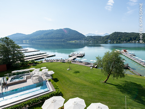 Ein malerischer Blick auf den See mit grünem Rasen, Poolbereich mit Liegestühlen und Sonnenschirmen, einem Steg mit Booten und baumbedeckten Hügeln im Hintergrund unter einem klaren blauen Himmel.
