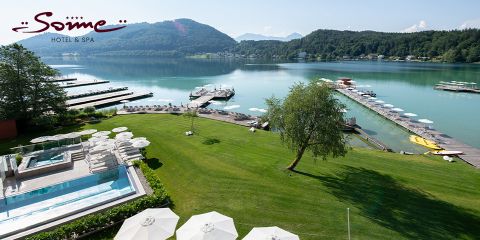 Ein malerisches Resort am Seeufer mit einem Hotellogo, Außenpools, grünen Rasenflächen, weißen Sonnenschirmen, Anlegestellen mit Booten, ruhigem Wasser und bewaldeten Hügeln im Hintergrund unter einem klaren Himmel.