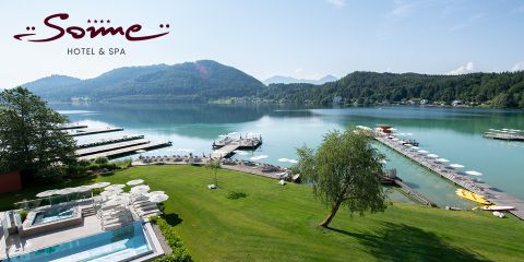 Ein malerisches Resort am Seeufer mit einem Hotellogo, Außenpools, grünen Rasenflächen, weißen Sonnenschirmen, Anlegestellen mit Booten, ruhigem Wasser und bewaldeten Hügeln im Hintergrund unter einem klaren Himmel.