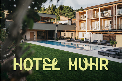 Ein modernes Hotel mit Holzbalkonen blickt auf einen Swimmingpool, der von Liegestühlen und grünem Gras umgeben ist, mit Bäumen im Hintergrund. Der Text "HOTEL MUHR" erscheint in großen gelben Buchstaben im Vordergrund.