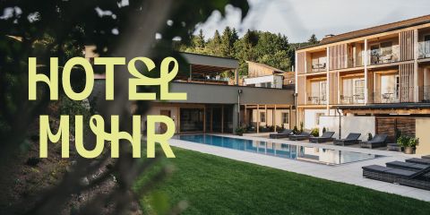 Ein modernes Hotel mit Balkonen und Blick auf einen Swimmingpool, umgeben von Liegestühlen und grünem Rasen. Ein großer Text auf der linken Seite lautet "HOTEL MUHR". Im Hintergrund sind Bäume und blauer Himmel zu sehen.