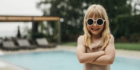 Lächelndes junges Mädchen mit langen blonden Haaren und weißer Sonnenbrille steht mit verschränkten Armen vor einem Schwimmbad an einem regnerischen Tag und trägt einen gestreiften Badeanzug. Im Hintergrund sind Liegestühle und Grünpflanzen zu sehen.