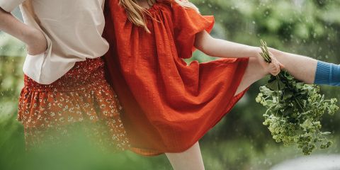Zwei eng beieinander stehende Personen, eine in einem weißen Oberteil und einem gemusterten roten Rock, die andere in einem roten Kleid mit hochgelegtem Bein, halten grüne Blätter in der Hand. Der Hintergrund ist grün und unscharf, was auf eine Umgebung im Freien hindeutet.