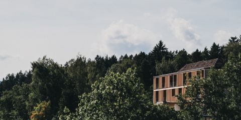 Ein modernes, hölzernes Gebäude, das teilweise von grünen Bäumen verdeckt wird, steht vor einem dichten Wald unter einem bewölkten Himmel.