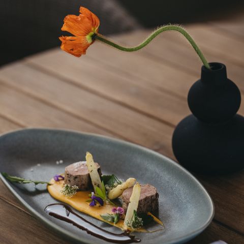 Ein modern gedeckter Tisch mit einem eleganten Gericht mit zwei Fleischstücken, weißem Spargel und bunten Beilagen auf einem grauen Teller, begleitet von einer orangefarbenen Blume in einer schwarzen Vase.