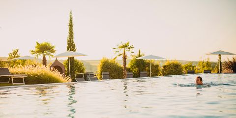 Eine Person schwimmt in einem großen Außenpool, umgeben von Palmen, Liegestühlen und Sonnenschirmen, mit einer malerischen Landschaft im Hintergrund unter warmem, goldenem Sonnenlicht.