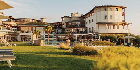 Ein modernes, mehrstöckiges Resort mit großen Fenstern und Balkonen bietet an sonnigen Tagen einen Blick auf eine Rasenfläche mit Palmen, Liegestühlen und einem Außenpool.