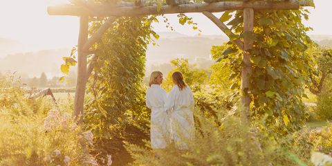 Zwei Frauen in weißen Gewändern stehen unter einem hölzernen Gartenbogen, der mit grünen Ranken bedeckt ist, umgeben von üppigen Pflanzen und warmem Sonnenlicht und genießen die friedliche Atmosphäre im Freien.