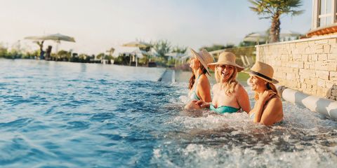 Vier Frauen mit Sonnenhüten entspannen sich und lächeln, während sie am Rande eines Außenpools sitzen, mit Palmen, Sonnenschirmen und Liegestühlen im Hintergrund an einem sonnigen Tag.