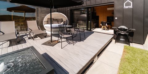 Eine moderne Außenterrasse mit Liegestühlen, einer runden Liege, einem Tisch mit Stühlen, einem schwarzen Sonnenschirm und einem Grill, neben einer schwarzen Hütte mit großen Glasschiebetüren. Im Vordergrund sind Gras und Steinfliesen zu sehen.