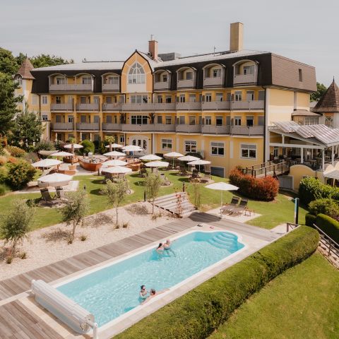 Ein großes gelbes Hotel mit weißer Verkleidung hat Balkone mit Blick auf einen angelegten Garten mit Liegestühlen, Sonnenschirmen und einem rechteckigen Außenpool, in dem Menschen schwimmen. Die Szene ist hell und sonnig.